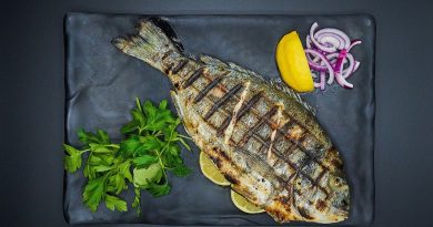 A grilled whole fish laying on a plate along with fresh herbs, onions and lemon.