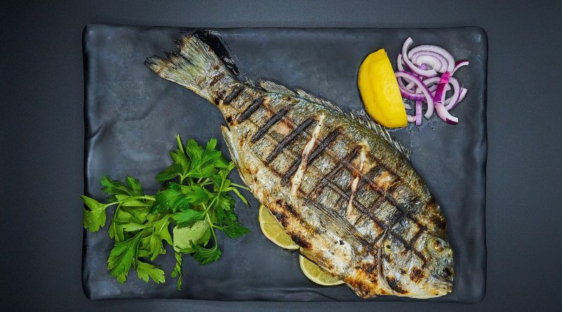 A grilled whole fish laying on a plate along with fresh herbs, onions and lemon.