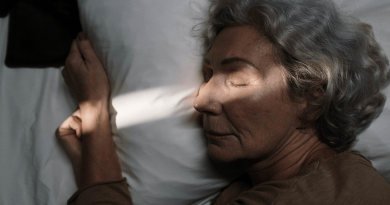 An older adult asleep in bed. A ray of light goes across her face and arm.