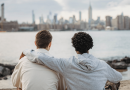 Two friends sitting close together, one with an arm over the other's shoulder. The pair is seen from behind as they look out over water with a city in the background.