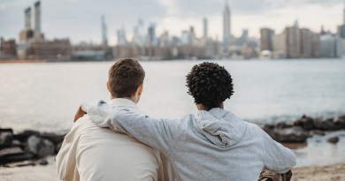 Two friends sitting close together, one with an arm over the other's shoulder. The pair is seen from behind as they look out over water with a city in the background.