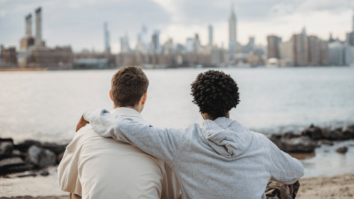 Two friends sitting close together, one with an arm over the other's shoulder. The pair is seen from behind as they look out over water with a city in the background.