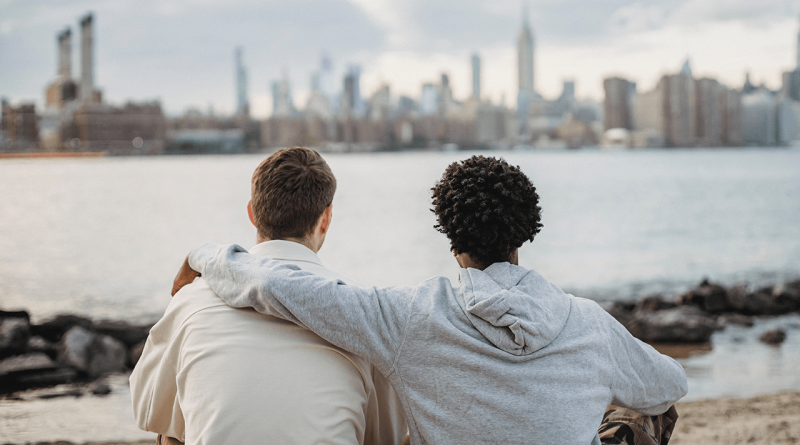 Two friends sitting close together, one with an arm over the other's shoulder. The pair is seen from behind as they look out over water with a city in the background.