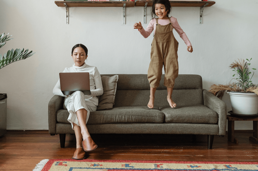 A parent using a laptop while their child jumps on the same sofa.