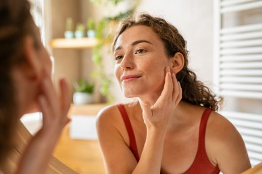 Beauty girl looking at mirror while touching her face and checking pimple, wrinkles and bags under the eyes, during morning beauty routine.