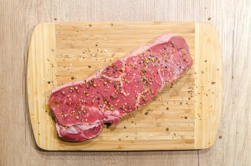 A strip of uncooked beef steak seasoned with peppercorn on a cutting board.