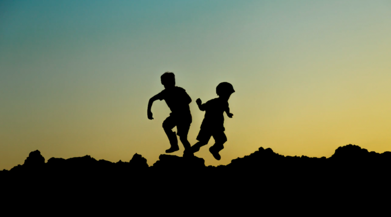 Two children playing on a rocky hilltop, silhouetted by the sun setting behind them