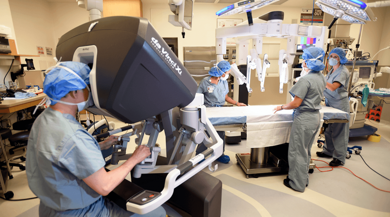 A surgeon sitting at a robotic surgery unit near an empty surgical bed.