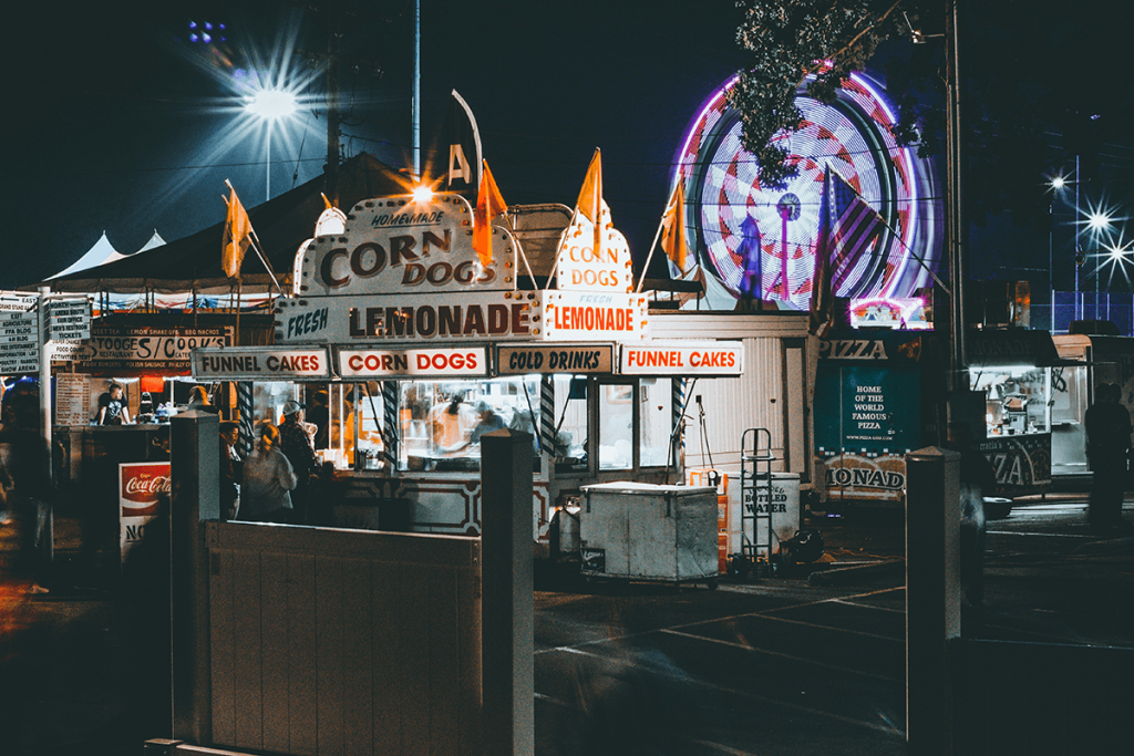 A brightly-lit corn dog stand at a carnival at night.