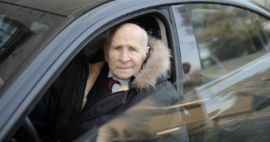 Older white man sitting in the driver's seat of a sedan. The car door is open.