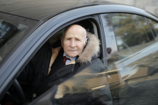 Older white man sitting in the driver's seat of a sedan. The car door is open.