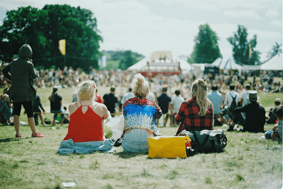 Three friends sitting in a field overlooking an outdoor festival.