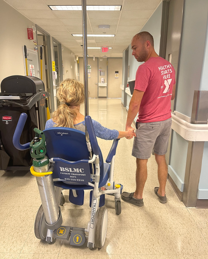 Allison using a wheelchair and holding her husband's hand as they walk through a hospital.