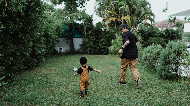 An adult and child kicking a soccer ball around in a green, lush backyard.
