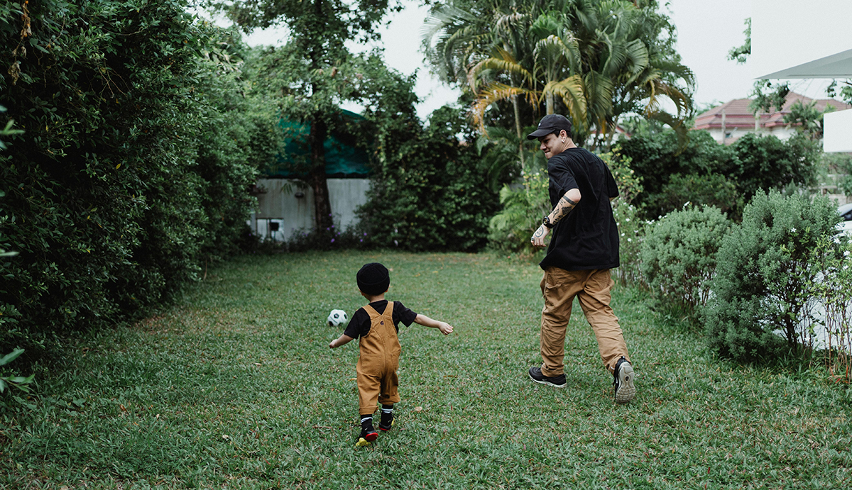 An adult and child kicking a soccer ball around in a green, lush backyard.