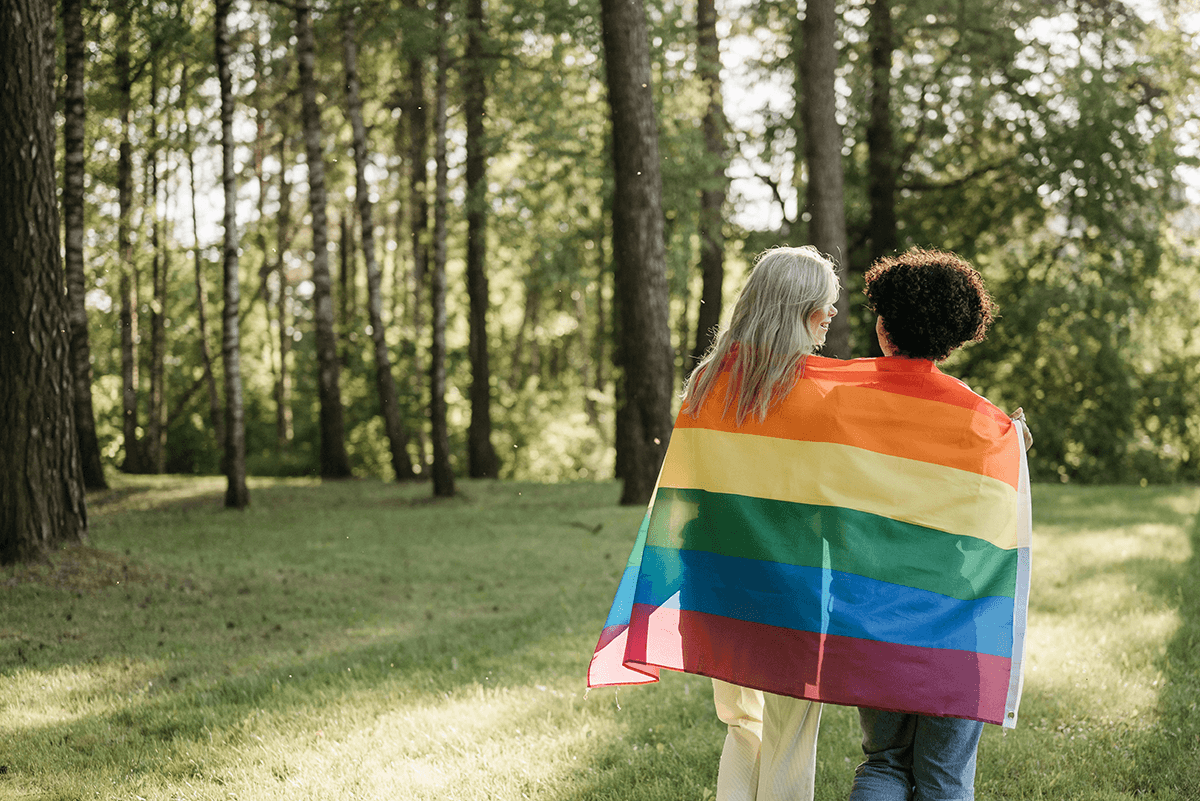 A pair of older adults walking through the woods, wrapped closely together in a Pride flag