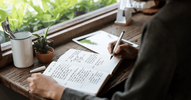 A person writing in their journal next to a window filled with greenery.