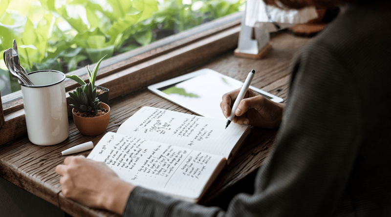 A person writing in their journal next to a window filled with greenery.