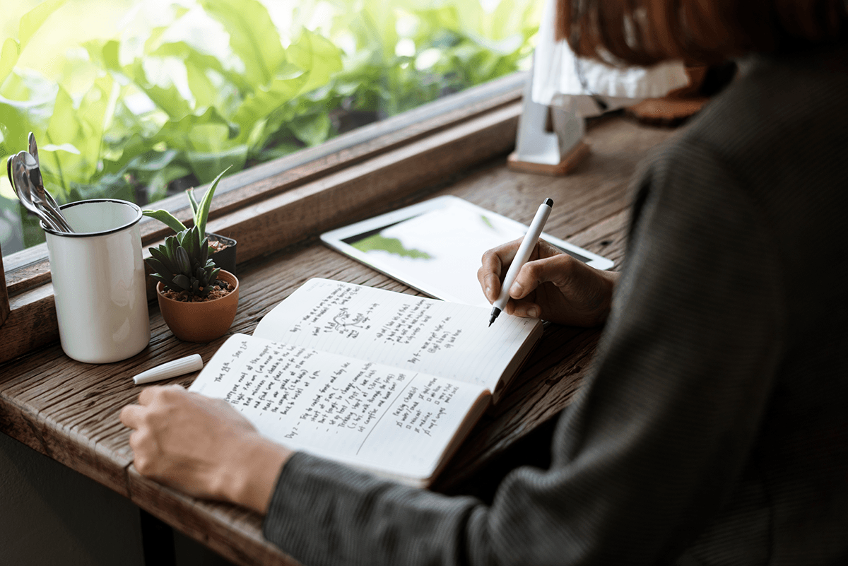 A person writing in their journal next to a window filled with greenery.