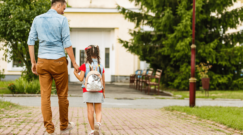 A caretaker and child walking toward a school, holding hands.