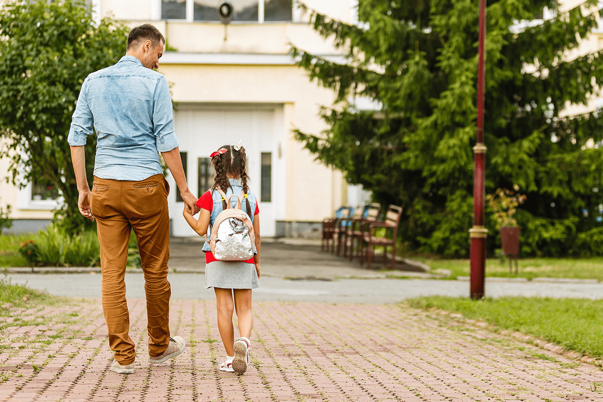 A caretaker and child walking toward a school, holding hands.