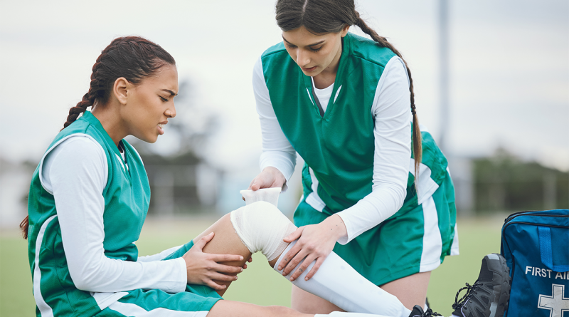 A soccer player helping another tape an injured knee on the field.