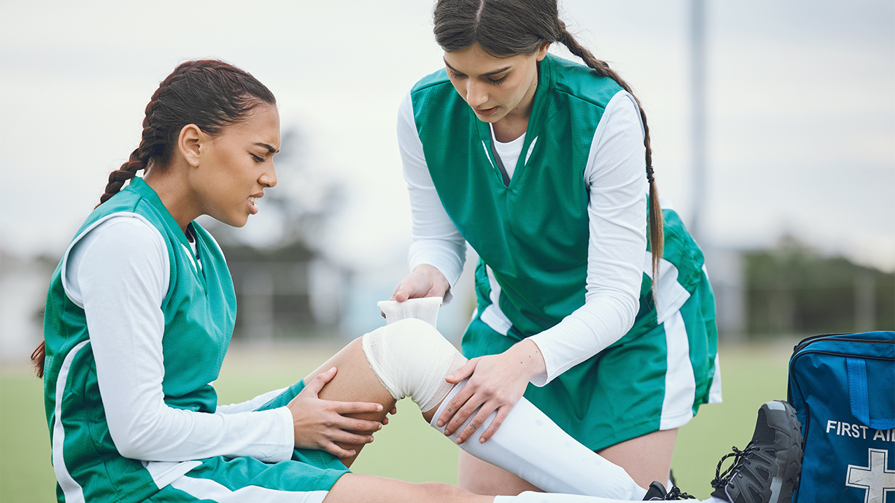 A soccer player helping another tape an injured knee on the field. 