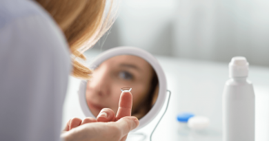 A person seen from behind. Their eye is visible in a mirror, and they're reaching a contact lens up to their eye.