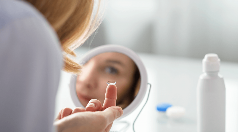 A person seen from behind. Their eye is visible in a mirror, and they're reaching a contact lens up to their eye.