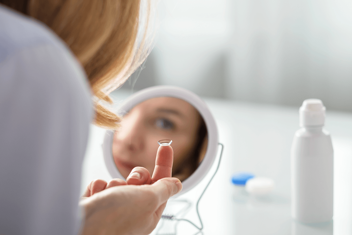 A person seen from behind. Their eye is visible in a mirror, and they're reaching a contact lens up to their eye.