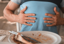 A bloated person holding their upper stomach with an empty plate of food in front of them.