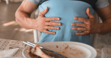 A bloated person holding their upper stomach with an empty plate of food in front of them.