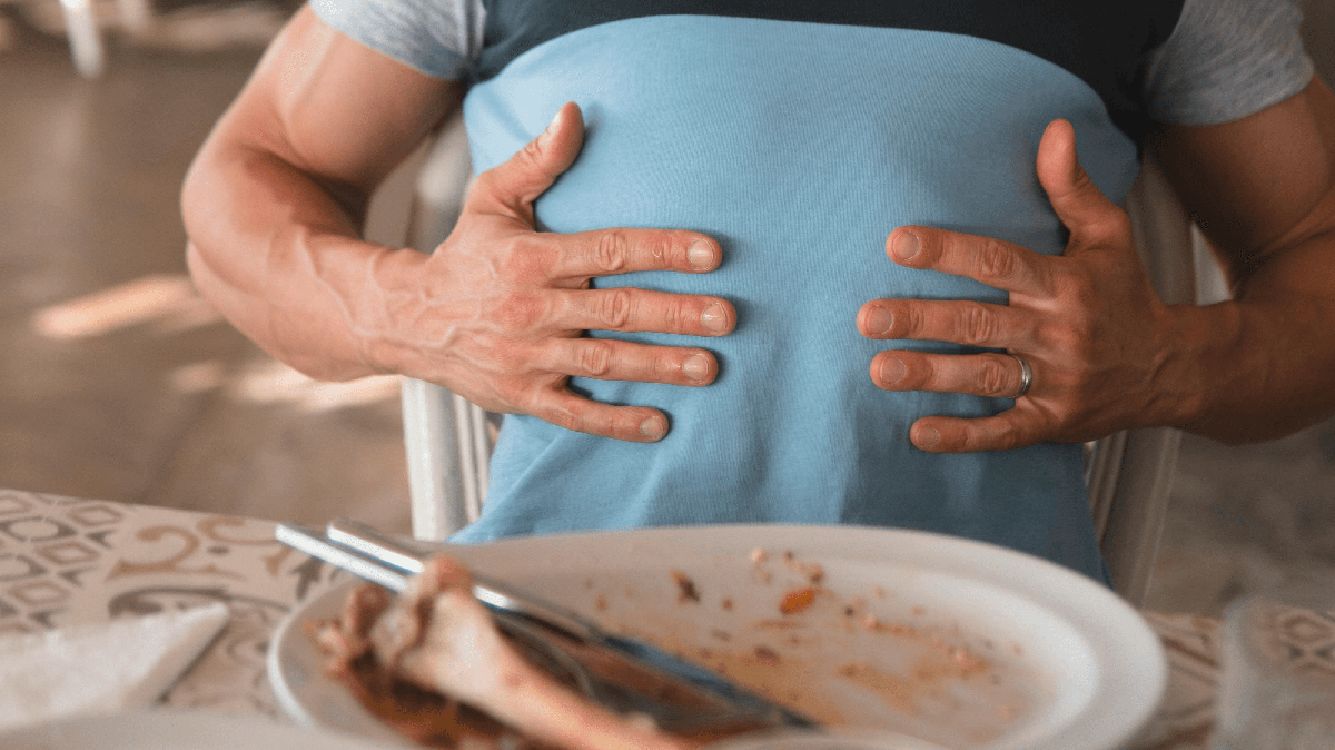 A bloated person holding their upper stomach with an empty plate of food in front of them.