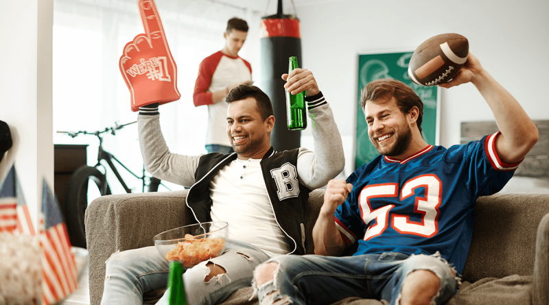 Two football fans on a coach cheering while a third snacks in the background.