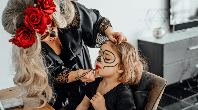 An adult using costume makeup to paint a skull face onto a child.