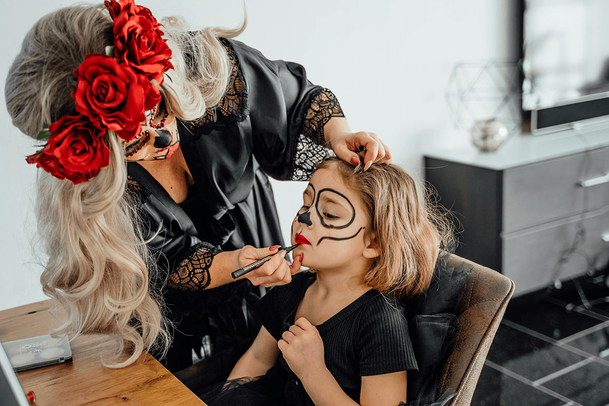 An adult using costume makeup to paint a skull face onto a child.