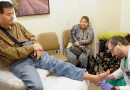 In an exam room, a doctor is kneeling to inspect a patient's propped up foot. The patient has his left arm in a cast, and another adult looks on.