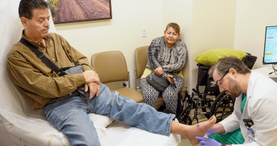 In an exam room, a doctor is kneeling to inspect a patient's propped up foot. The patient has his left arm in a cast, and another adult looks on.