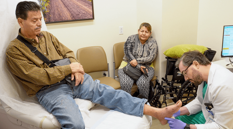 In an exam room, a doctor is kneeling to inspect a patient's propped up foot. The patient has his left arm in a cast, and another adult looks on.