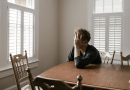 A person sitting at a kitchen table with their head in their hands, appearing sad.