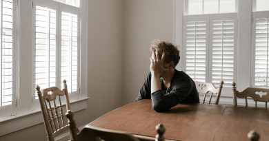 A person sitting at a kitchen table with their head in their hands, appearing sad.