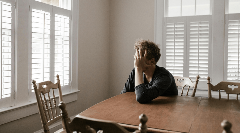 A person sitting at a kitchen table with their head in their hands, appearing sad.