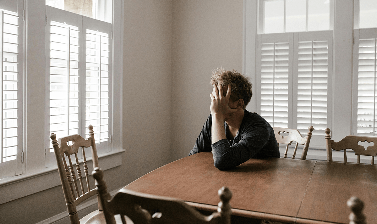 A person sitting at a kitchen table with their head in their hands, appearing sad.