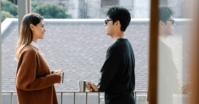 A pair of siblings holding metal coffee mugs standing on a balcony having a conversation. Their mood appears happy, but also serious.