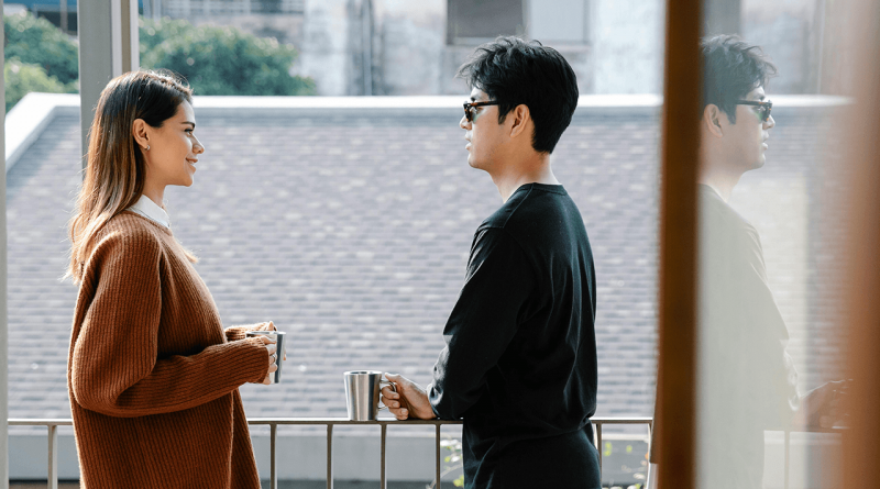 A pair of siblings holding metal coffee mugs standing on a balcony having a conversation. Their mood appears happy, but also serious.