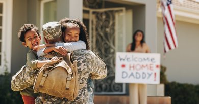 Two children hugging their father, who is dressed in a military uniform. His wife holds a sign reading "Welcome home daddy!" in the background.