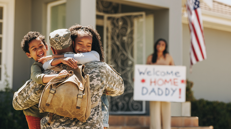 Two children hugging their father, who is dressed in a military uniform. His wife holds a sign reading "Welcome home daddy!" in the background.