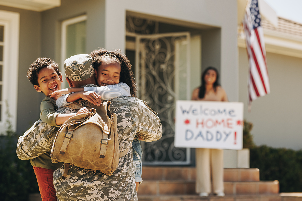 Two children hugging their father, who is dressed in a military uniform. His wife holds a sign reading "Welcome home daddy!" in the background.