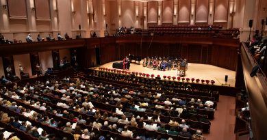 A brown-tinted performance hall with a large seated crowd watching students on the stage during a graduation.