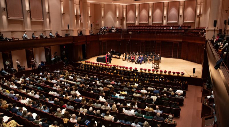 A brown-tinted performance hall with a large seated crowd watching students on the stage during a graduation.